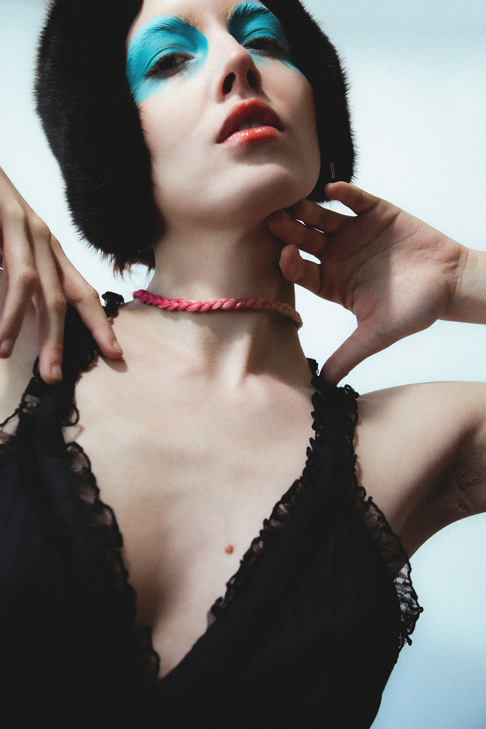 Close up portrait of Autry with bright blue makeup on wearing a fuzzy black hat, posing with their hands by their face